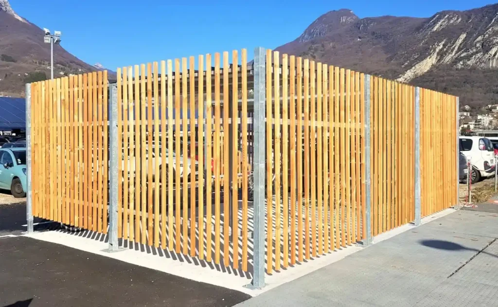 Abri vélo ByCommute en bois avec structure en lattes verticales, installé sur un parking avec montagnes en arrière-plan.