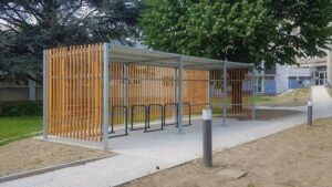 Abri vélo ouvert ByCommute avec lattes de bois verticales, situé dans un espace vert près d'un bâtiment.