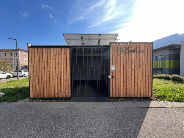 Un abri vélo fermé avec des parois en bois, équipé d’une porte métallique sécurisée et de panneaux solaires sur le toit, installé dans un environnement urbain avec des bâtiments modernes et un paysage montagnard en arrière-plan.