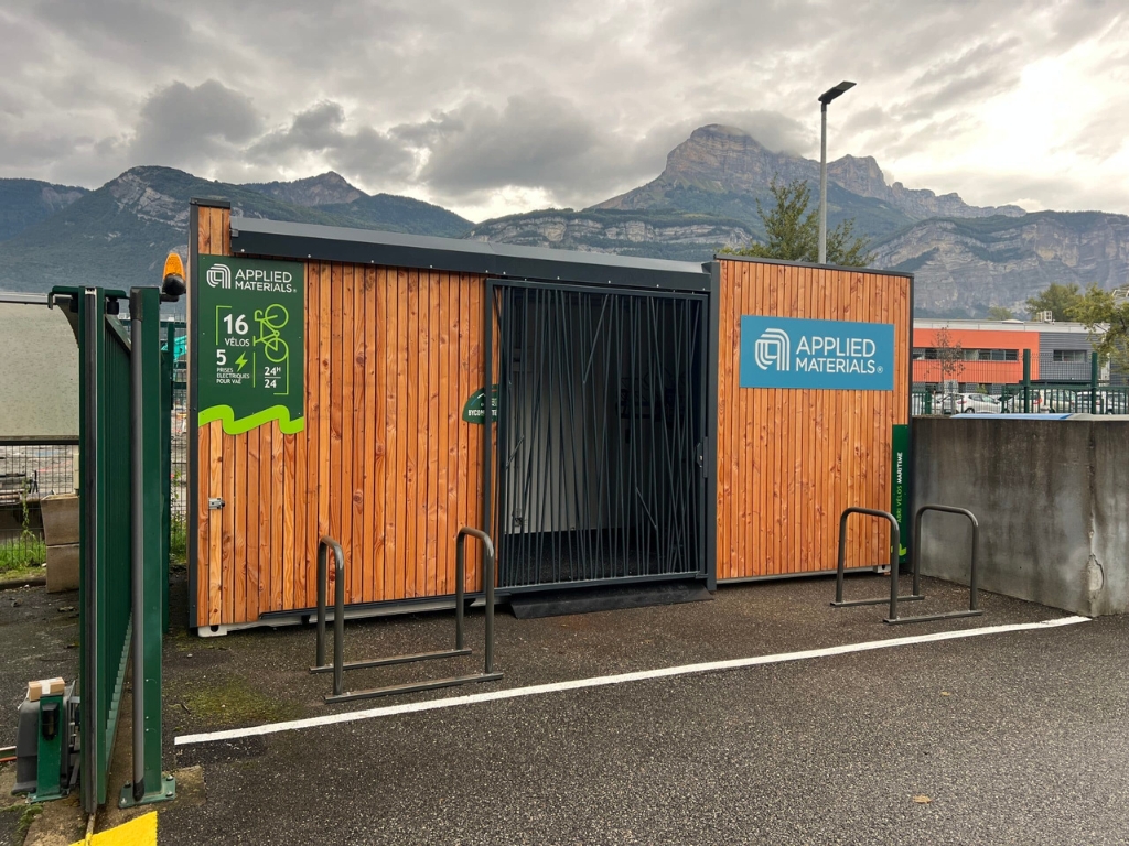 Abri vélo container en bois avec logo Applied Materials et Bycommute, situé dans un parking avec des supports pour vélos et des montagnes visibles en arrière-plan.