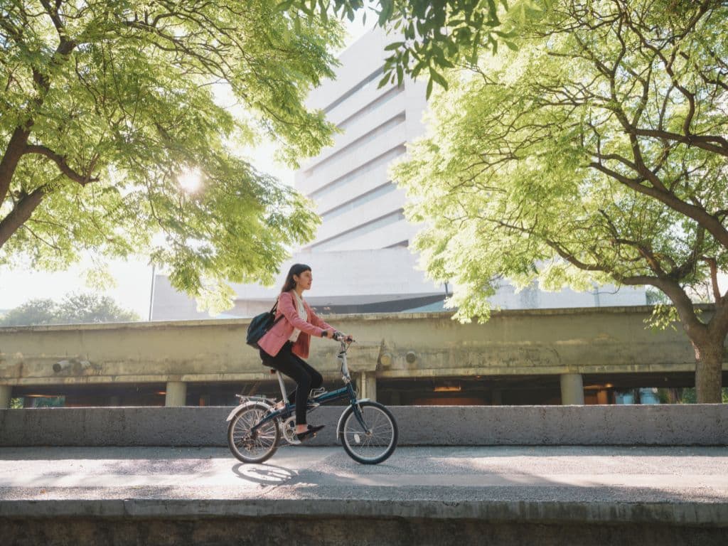 Femme à vélo en ville, circulant sur une piste cyclable ombragée par des arbres, avec un sac à dos.