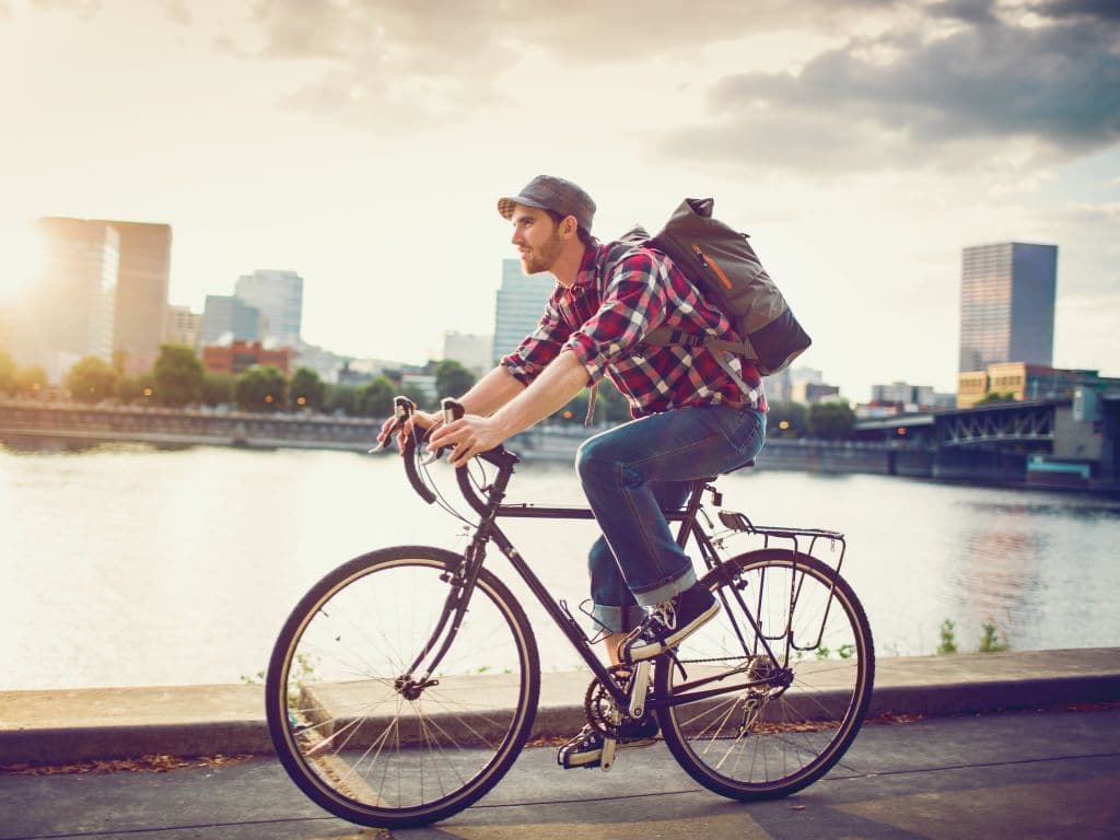 Homme à vélo en ville, portant un sac à dos, pédalant le long d'une rivière avec des immeubles en arrière-plan.