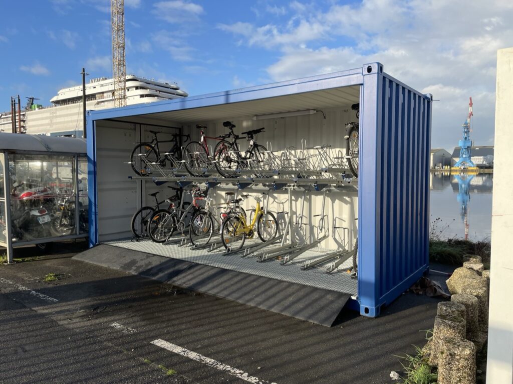 Abri vélo container ouvert bleu équipé de racks sur deux niveaux pour le stationnement de vélos, installé sur un parking urbain ou près d’un quai.