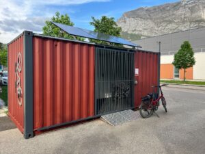 Un abri vélo container rouge avec une structure métallique sécurisée, équipé de panneaux solaires sur le toit, placé à proximité d’un bâtiment moderne avec des montagnes visibles en arrière-plan.