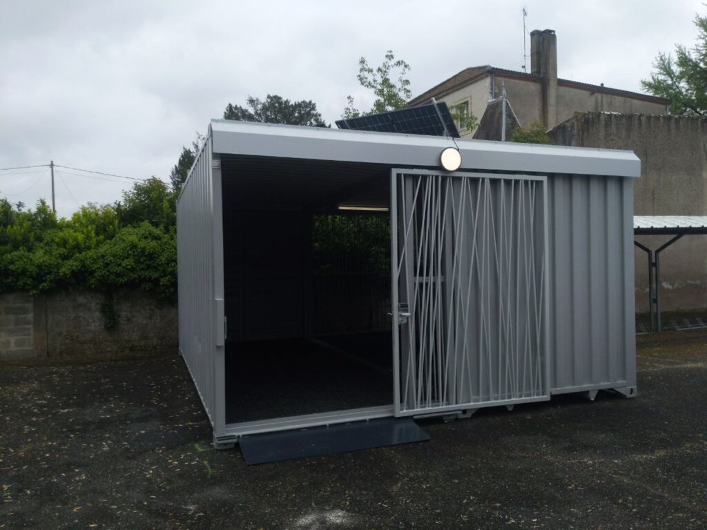 Abri vélo container fermé avec racks de rangement intérieurs, équipé d'une porte sécurisée et d'un panneau solaire sur le toit, situé sur une aire extérieure.