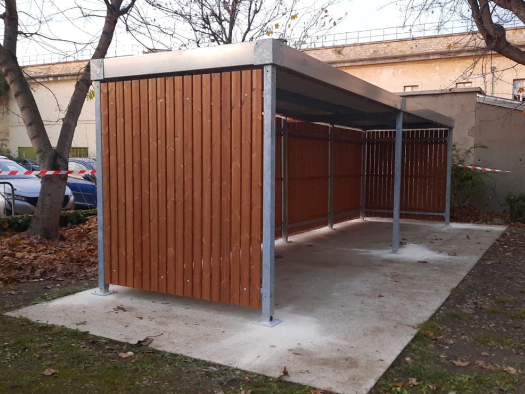 Abri vélo en bois avec structure métallique installé sur une dalle béton, entouré d'arbres dans une zone résidentielle.